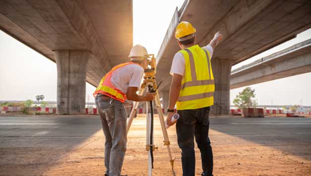 Two construction workers stand in front of a survey laser