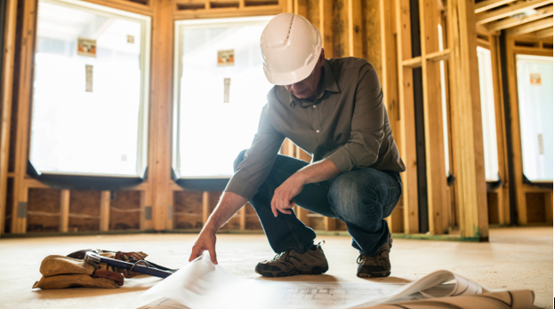 A contractor wearing a hardhat kneels to examine blueprints that rest on the ground.