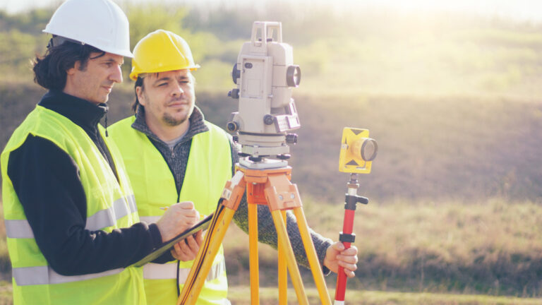 Two surveyors wearing high-vis vests observe a piece of survey equipment.