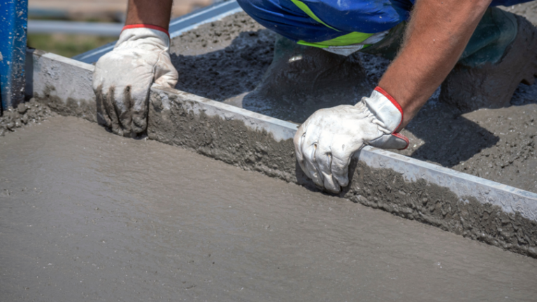 A concrete worker smooth's out a large concrete slab with a long flat tool.